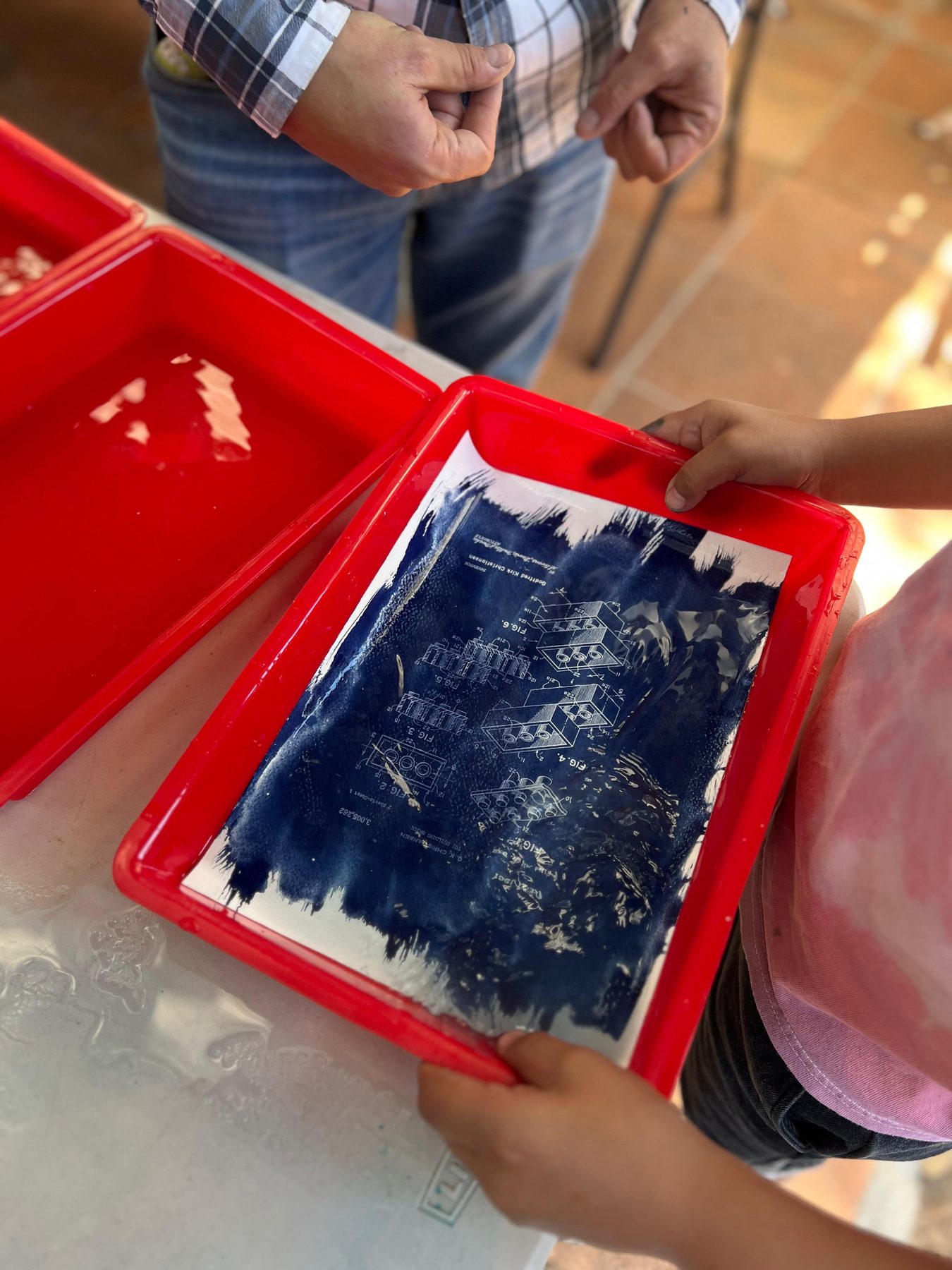 Manos sacando una lámina azul de cianotipia de una charola roja, en una Experiencia de Tribu