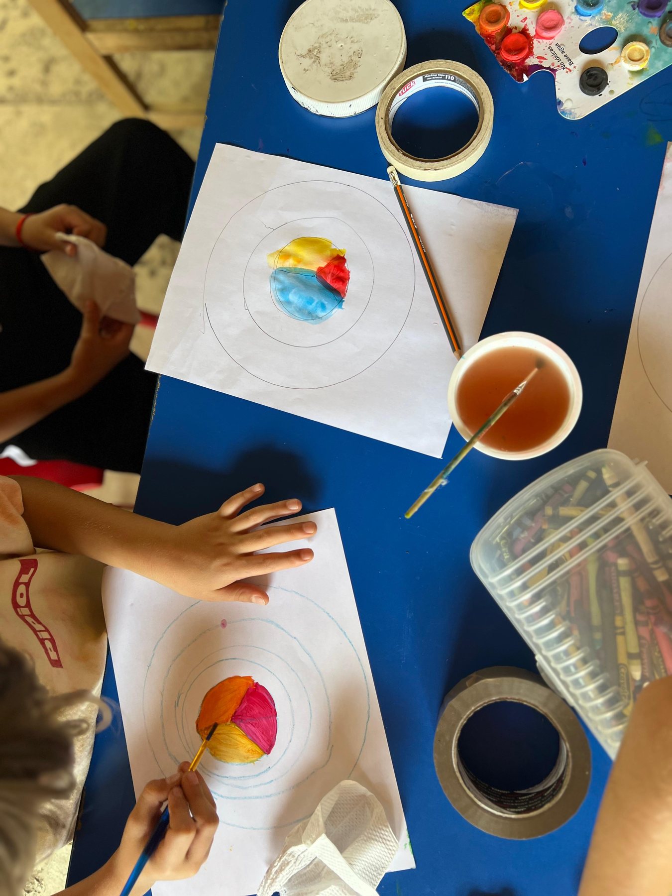 Manos de ninos pintando circulos con acuarela sobre una mesa azul, en una Experiencia de Tribu